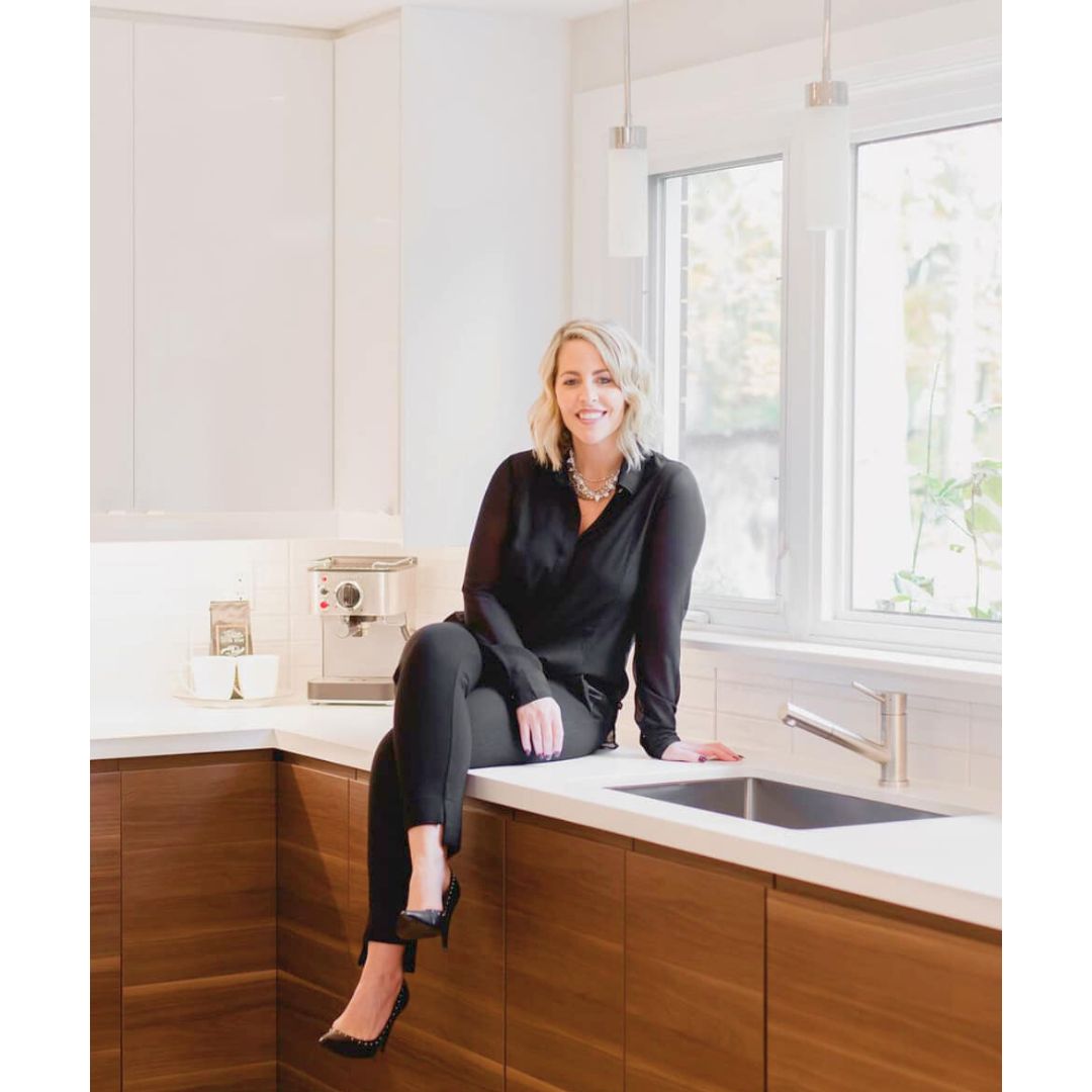 Woman sitting on white quartz countertop beside kitchen sink with natural lighting coming through windows.