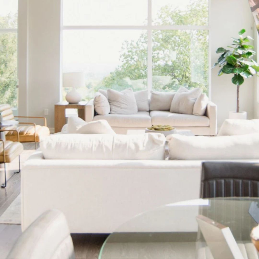 Stylish open-plan living area with white sofas, tan leather chairs, and a glass dining table. Large windows provide ample natural light, while a fiddle-leaf fig plant adds a pop of green.