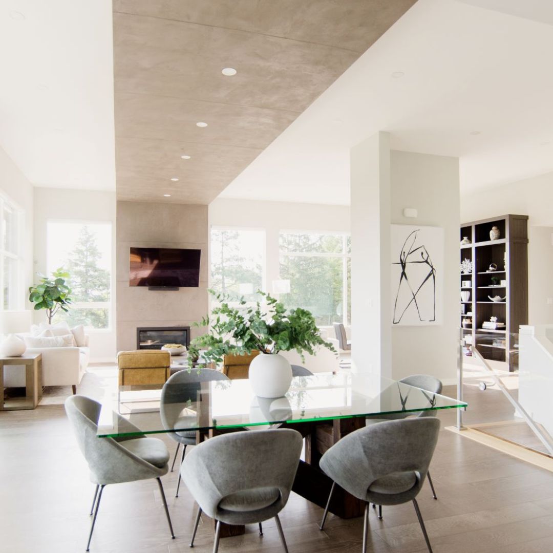 Contemporary dining area featuring a glass table with gray chairs, set against a backdrop of white walls and large windows. A striking concrete ceiling detail runs down the center, adding visual interest.