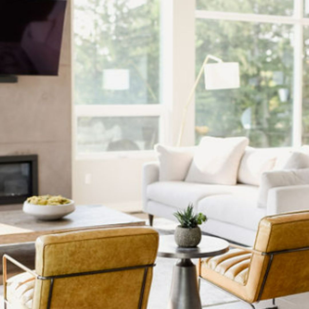 Bright modern living room with large windows, white walls, and wood floors. Features tan leather chairs, white sofa, wall-mounted TV, and fireplace. Natural light floods the space, highlighting the minimalist decor