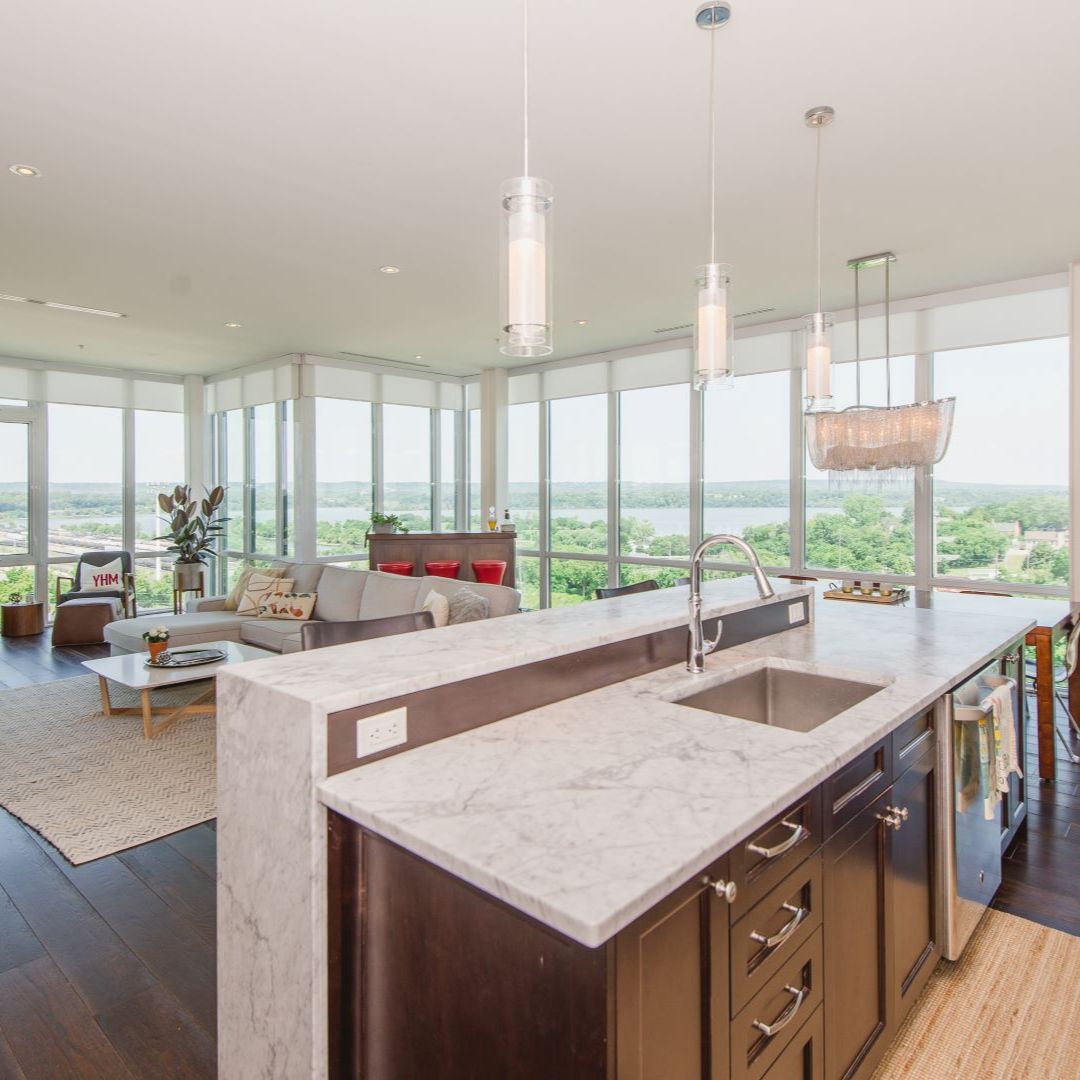 Luxurious open-concept apartment with stunning panoramic views. Modern kitchen featuring marble countertops and stainless steel appliances flows into a chic living area. Floor-to-ceiling windows showcase breathtaking vistas of lush greenery and water. Sleek pendant lights, hardwood floors, and contemporary furnishings