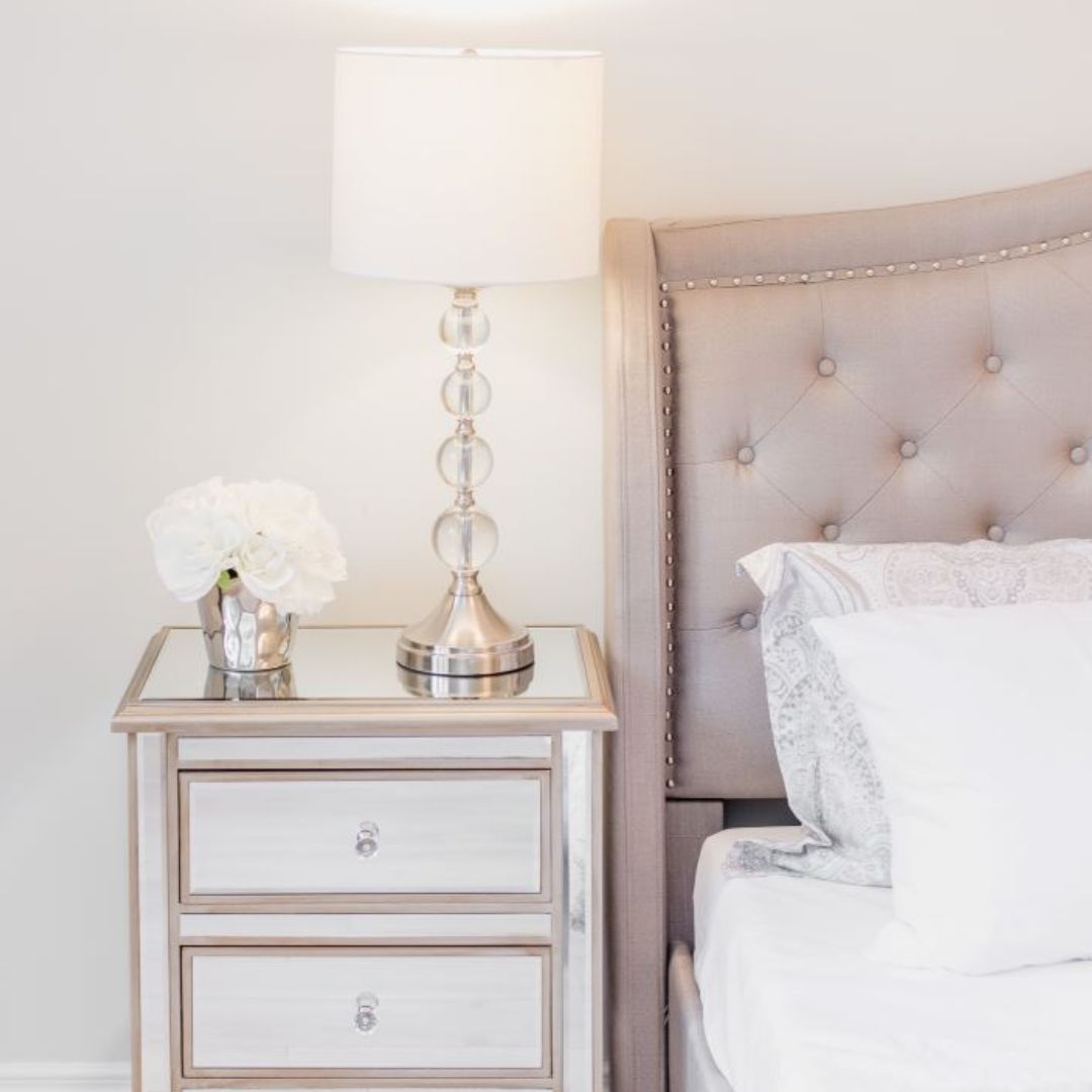 Close-up of chic bedside table with glass lamp, mirrored finish, and white flowers, next to tufted headboard with nailhead trim, highlighting bedroom styling details