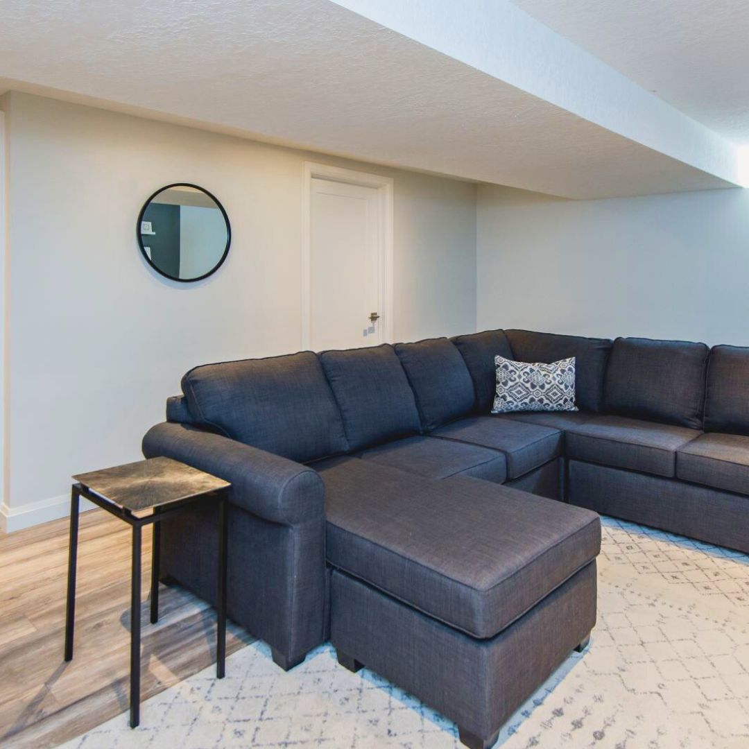 Spacious basement family room with grey sectional sofa, round mirror, and white walls creating a bright, inviting space.