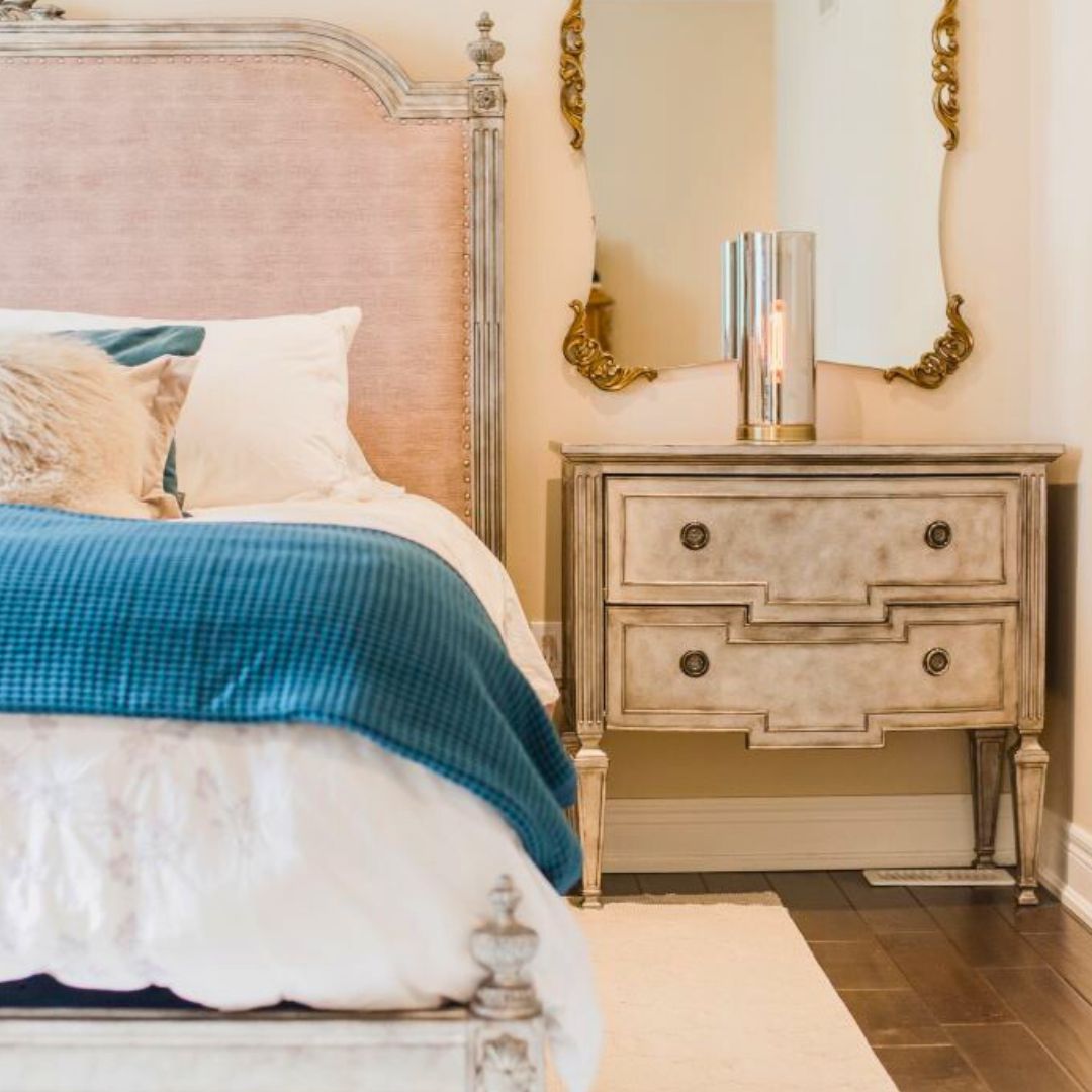Classic bedroom design featuring a pink upholstered headboard, vintage-inspired nightstand, and modern glass lamp. Blue throw and pillows contrast beautifully with the neutral color scheme.