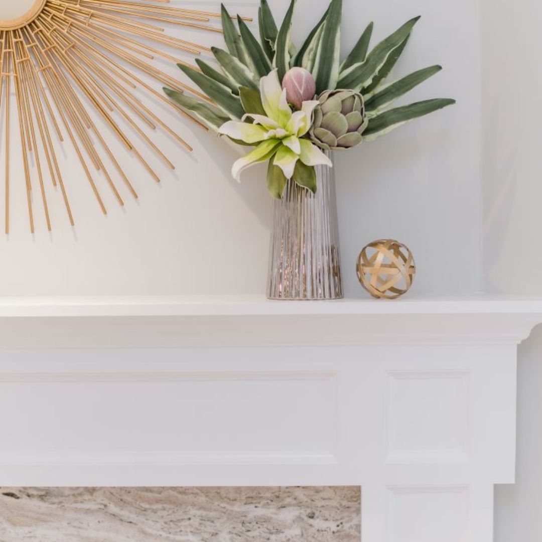 Coastal-inspired mantel decor featuring a gold sunburst mirror, silver vase with tropical plants, and decorative golden sphere. 
