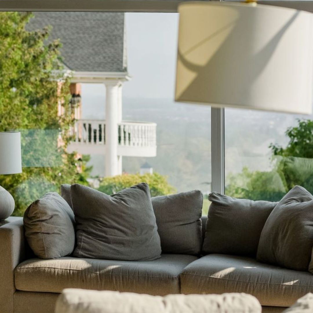 Cozy living room with a big gray couch and a view of the ocean. You can see trees and a white house outside. It looks like a fancy beach house where you'd love to relax