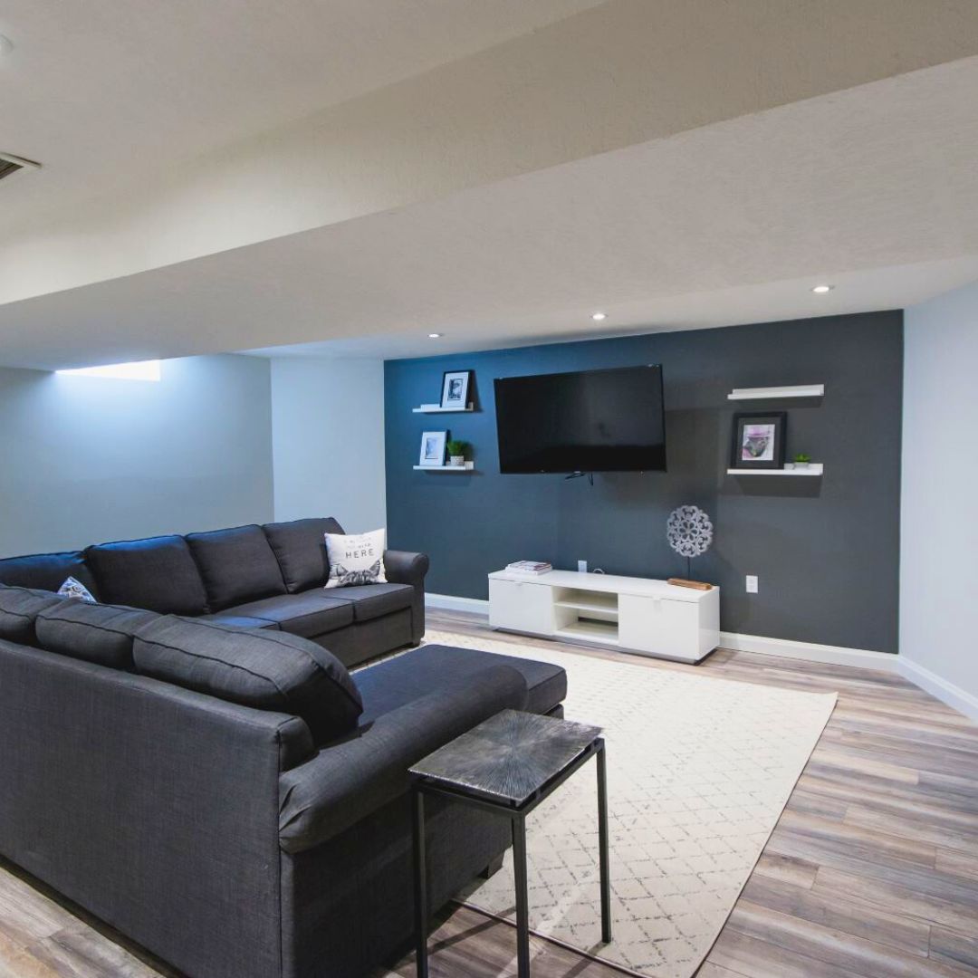 Cozy basement living room with dark blue accent wall, mounted TV, and large grey sectional sofa on light wood floors