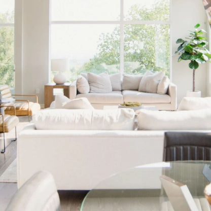 Stylish open-plan living area with white sofas, tan leather chairs, and a glass dining table. Large windows provide ample natural light, while a fiddle-leaf fig plant adds a pop of green.