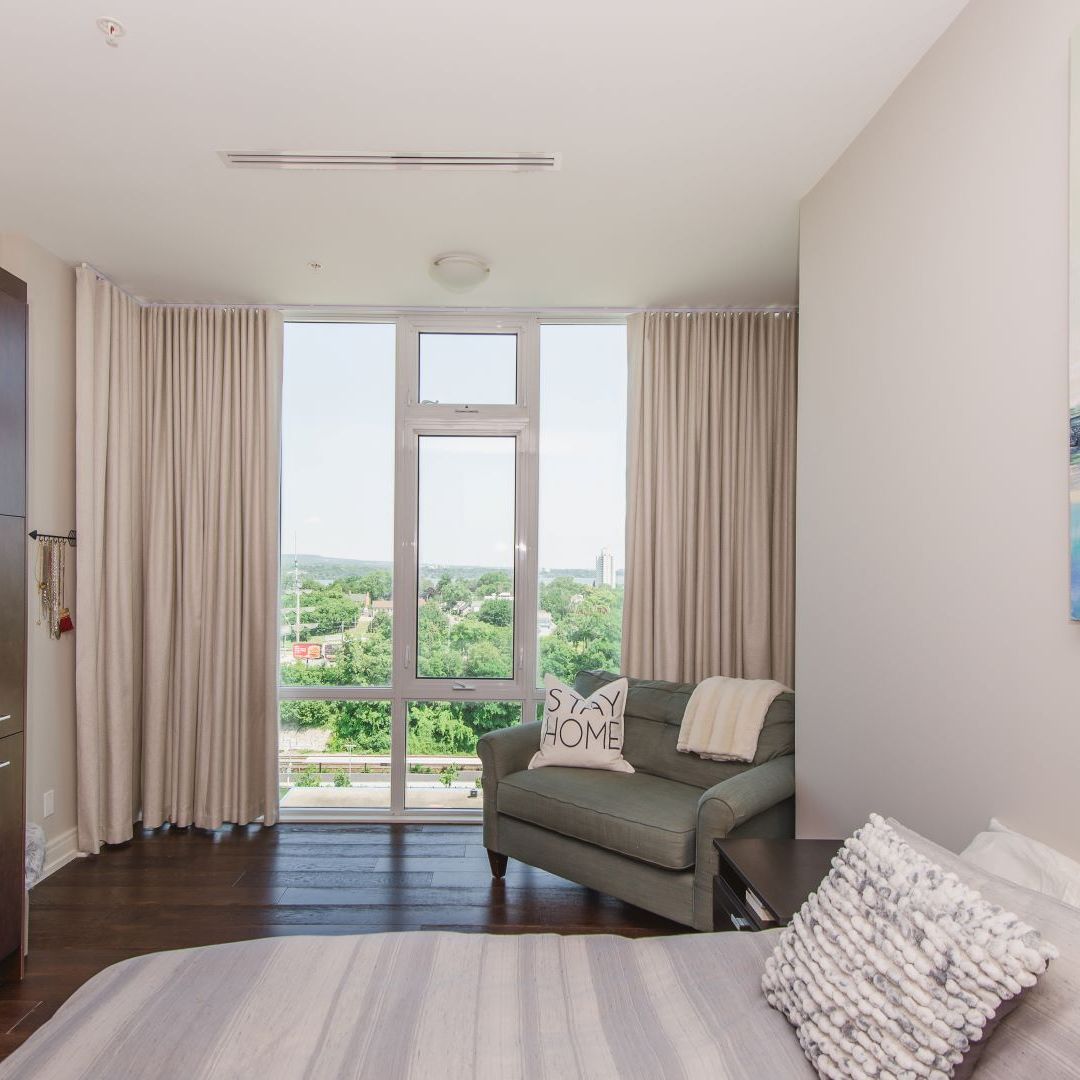 Cozy apartment bedroom with large windows offering a green city view, featuring a comfortable loveseat and "Stay Home" pillow.