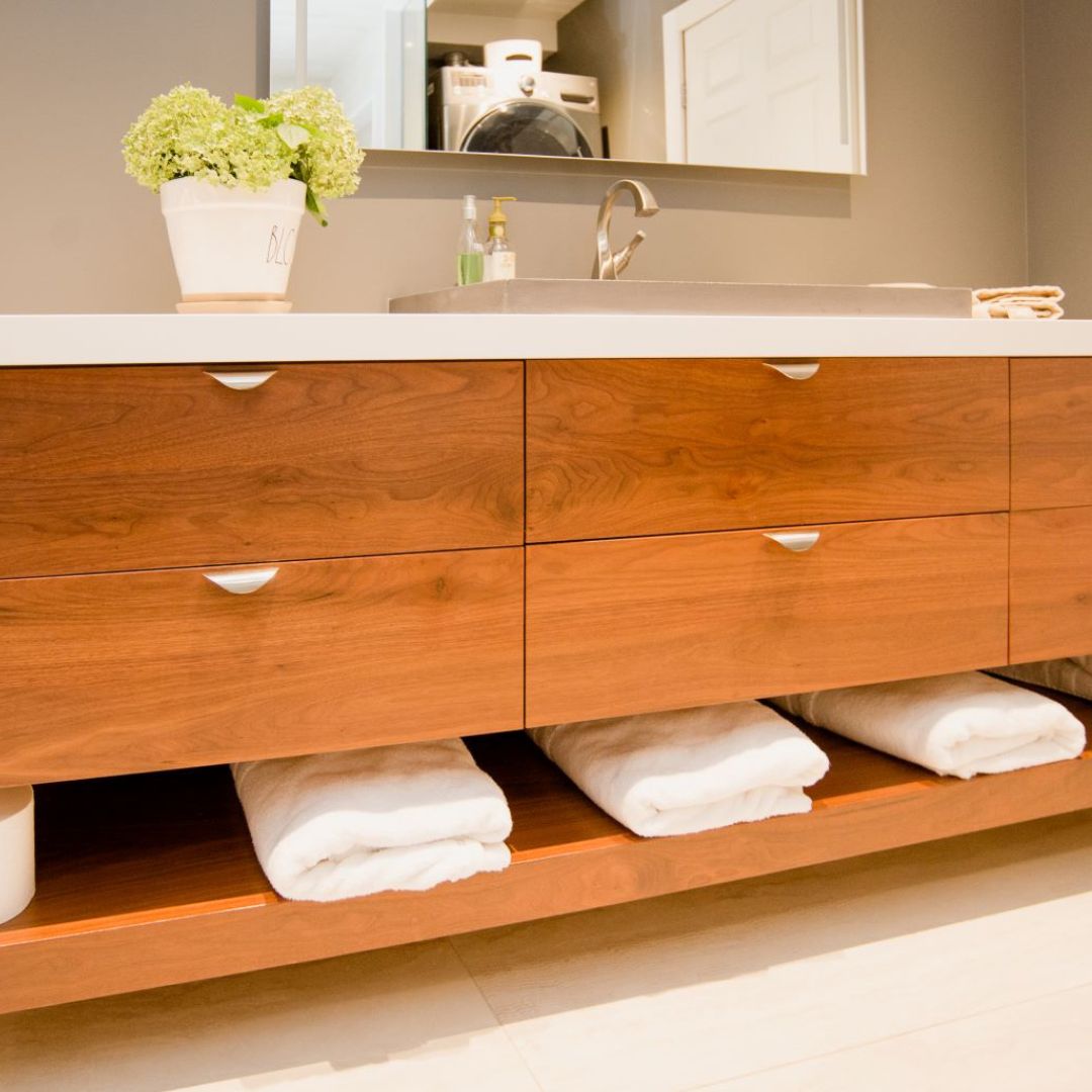 Expansive custom bathroom vanity featuring rich wood cabinetry, white quartz countertop, and open bottom shelving stocked with plush white towels. A stainless steel sink and modern faucet complement the design, while a mirror and glimpse of laundry appliances suggest a multifunctional space perfect for modern home organization.