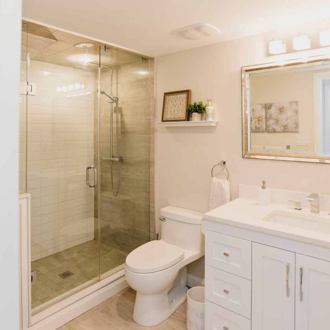 Bright bathroom with glass shower enclosure, white vanity, and large framed mirror. Beige tile flooring and walls create a warm, inviting atmosphere.