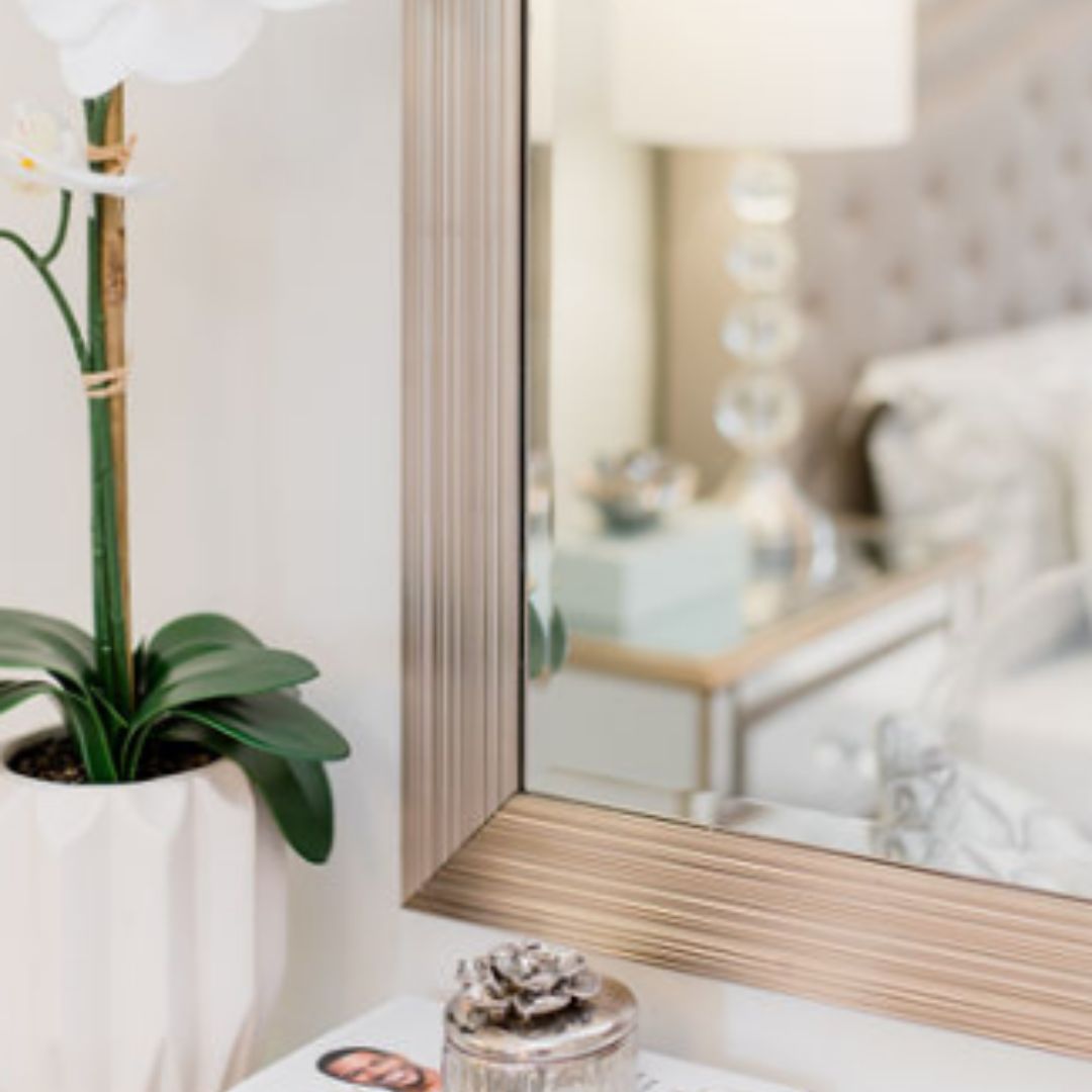 Bedroom corner with wood-framed mirror, white orchid, and glimpse of soft bedding, showing elegant interior design details.