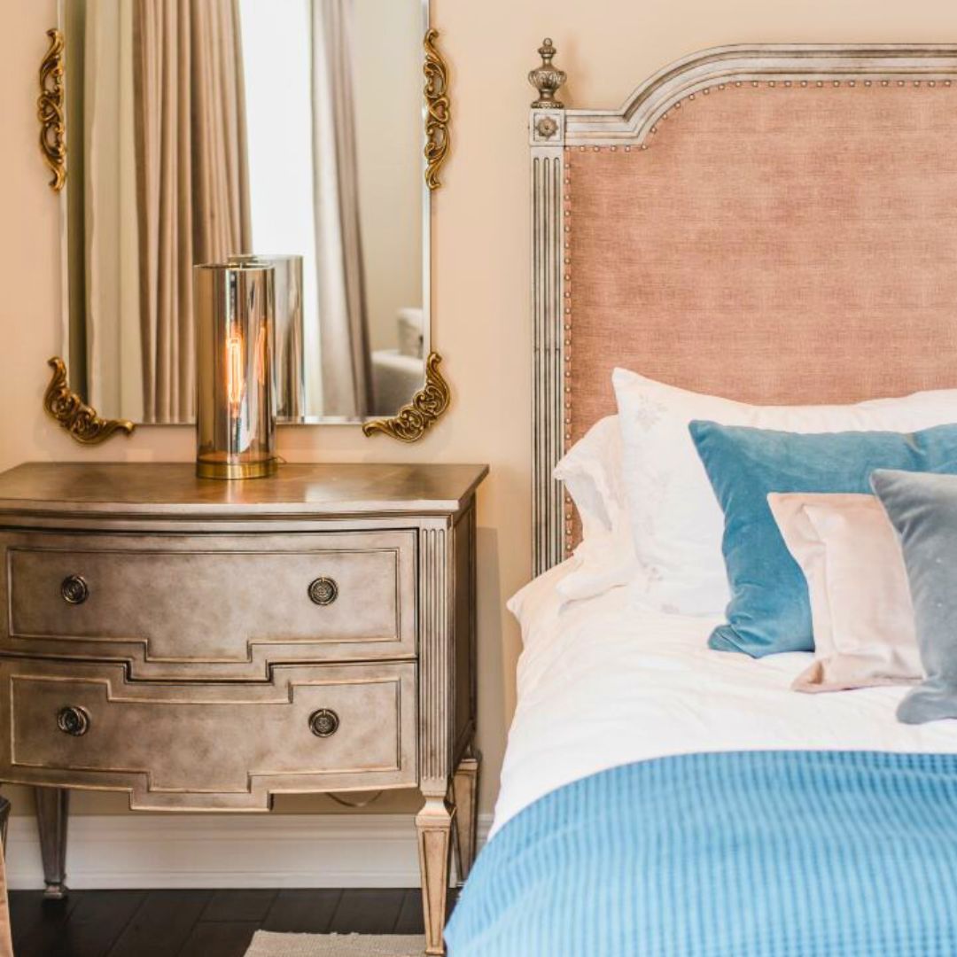 Elegant bedroom with blush headboard, antique-style nightstand, gold lamp, ornate mirror, and blue accent pillows