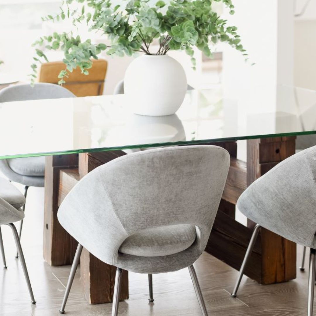 Elegant dining area with a glass-top table and modern gray chairs. The rustic wooden base contrasts with the sleek tabletop. A stunning white vase with cascading eucalyptus makes a beautiful centerpiece. Perfect for contemporary home decor, mixing different textures and materials.