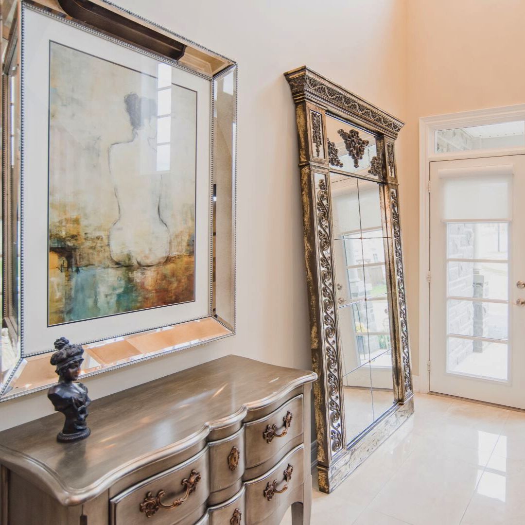 Sophisticated entryway with ornate gold mirror, silver chest of drawers, and framed abstract artwork. French doors and sheer curtains allow natural light to illuminate the elegant space.