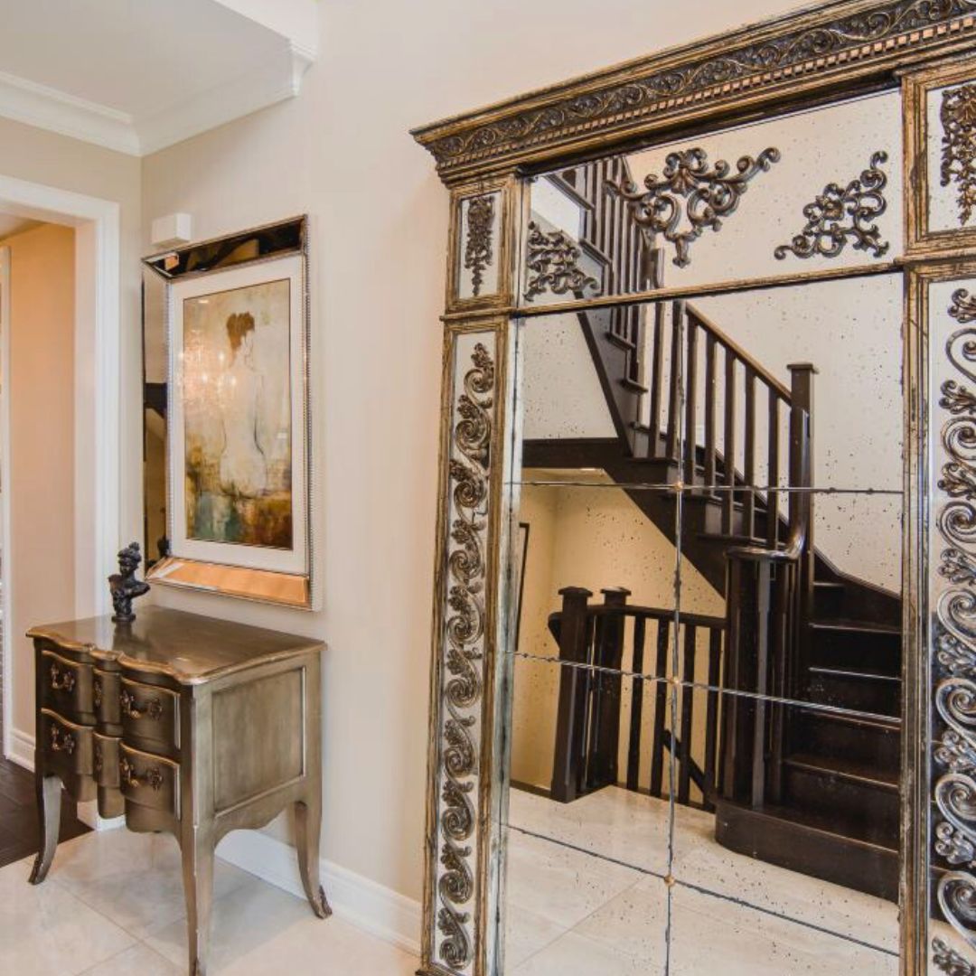 elegant entryway featuring a large ornate gold mirror, antique-style chest, and framed artwork. The mirror reflects a dark wood staircase, creating depth and elegance in the space.
