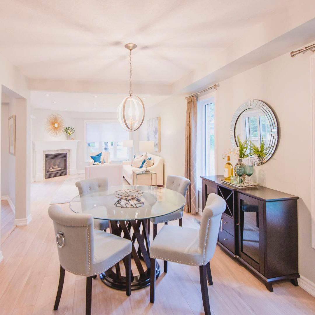 Stunning open-concept dining and living area with chic decor. Round glass table and plush gray chairs create an inviting dining space. Stylish orb chandelier hangs above. Dark wood sideboard with circular mirror adds elegance. Bright living room with fireplace visible in background. Light hardwood floors and soft colors throughout