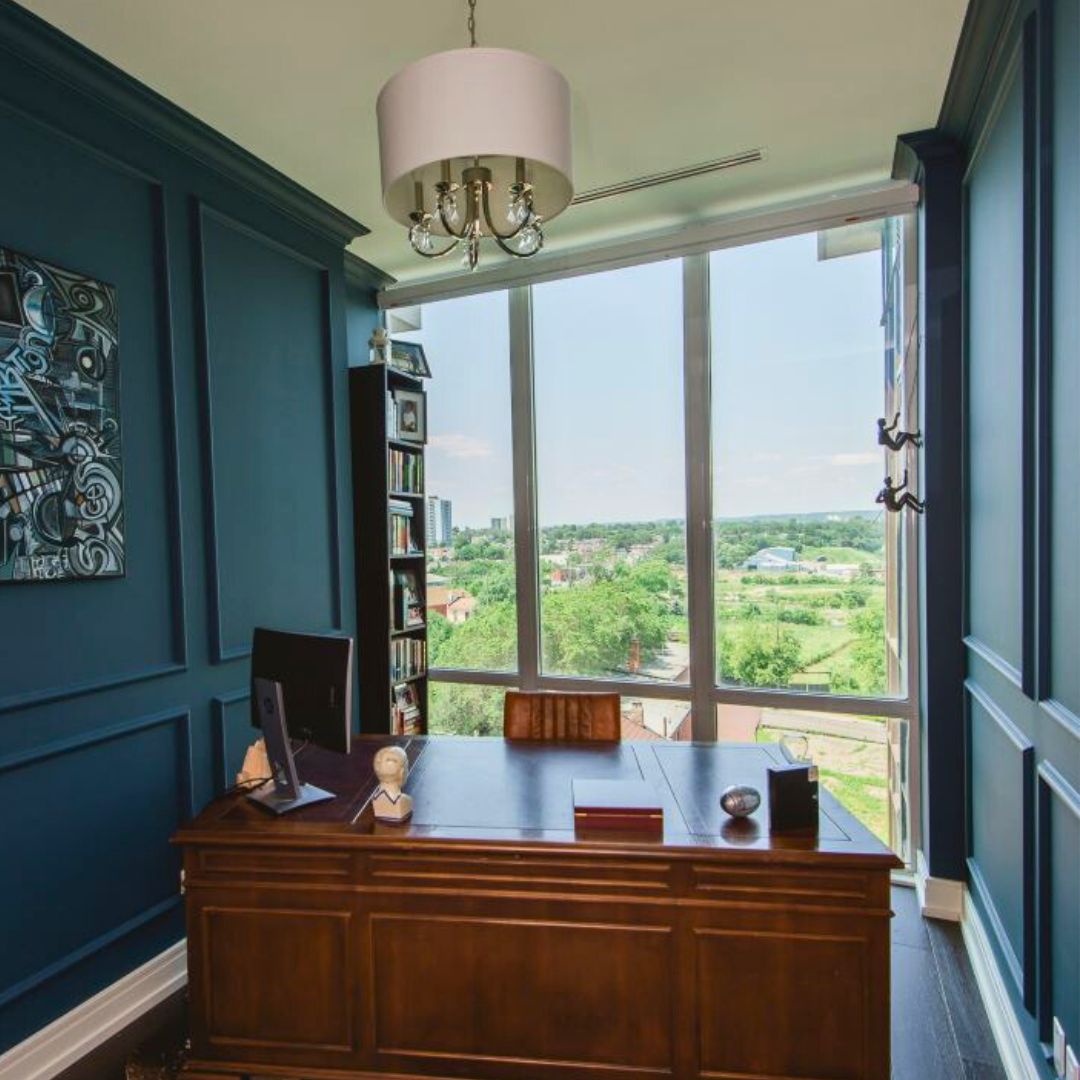 Executive home office design  featuring a large wooden desk, built-in bookcases, and floor-to-ceiling windows with scenic views. The space includes rich blue walls with panel molding and a modern chandelier