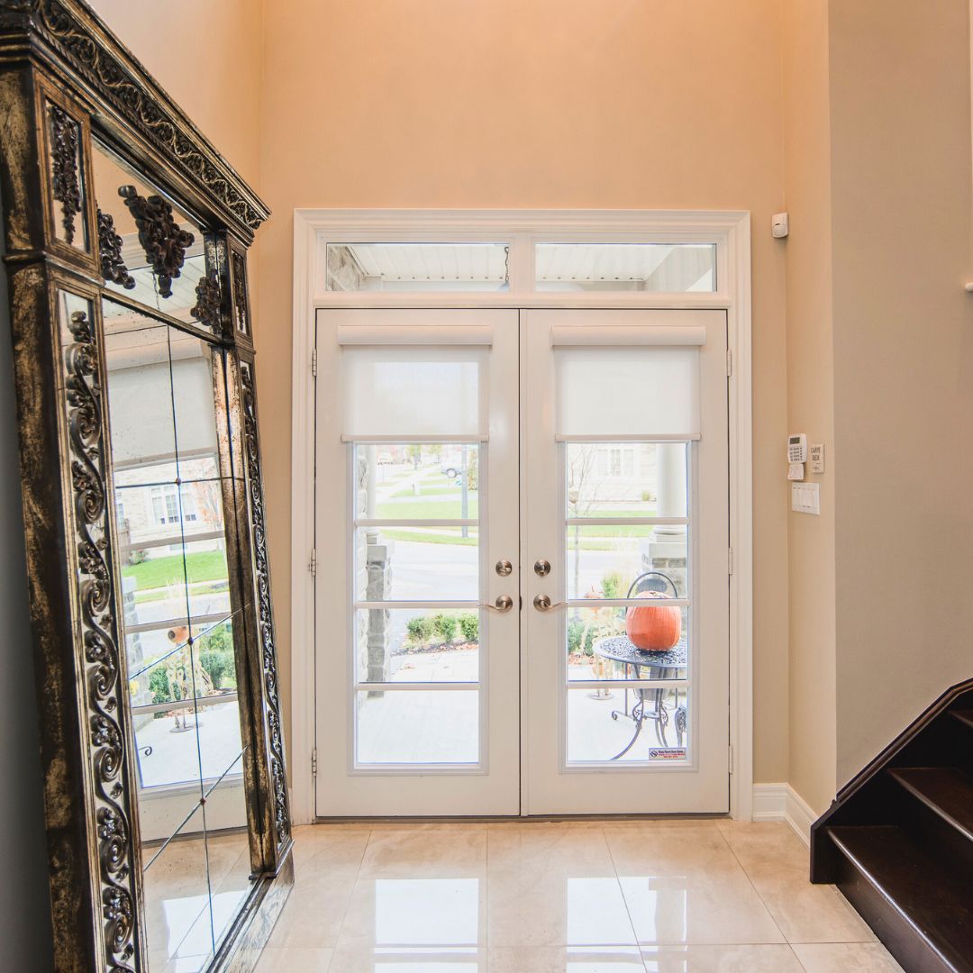 Grand entryway with ornate floor-to-ceiling mirror and white French doors. Polished tile floors reflect light, while peach walls create a warm, welcoming atmosphere. Glimpse of outdoor patio visible through doors.