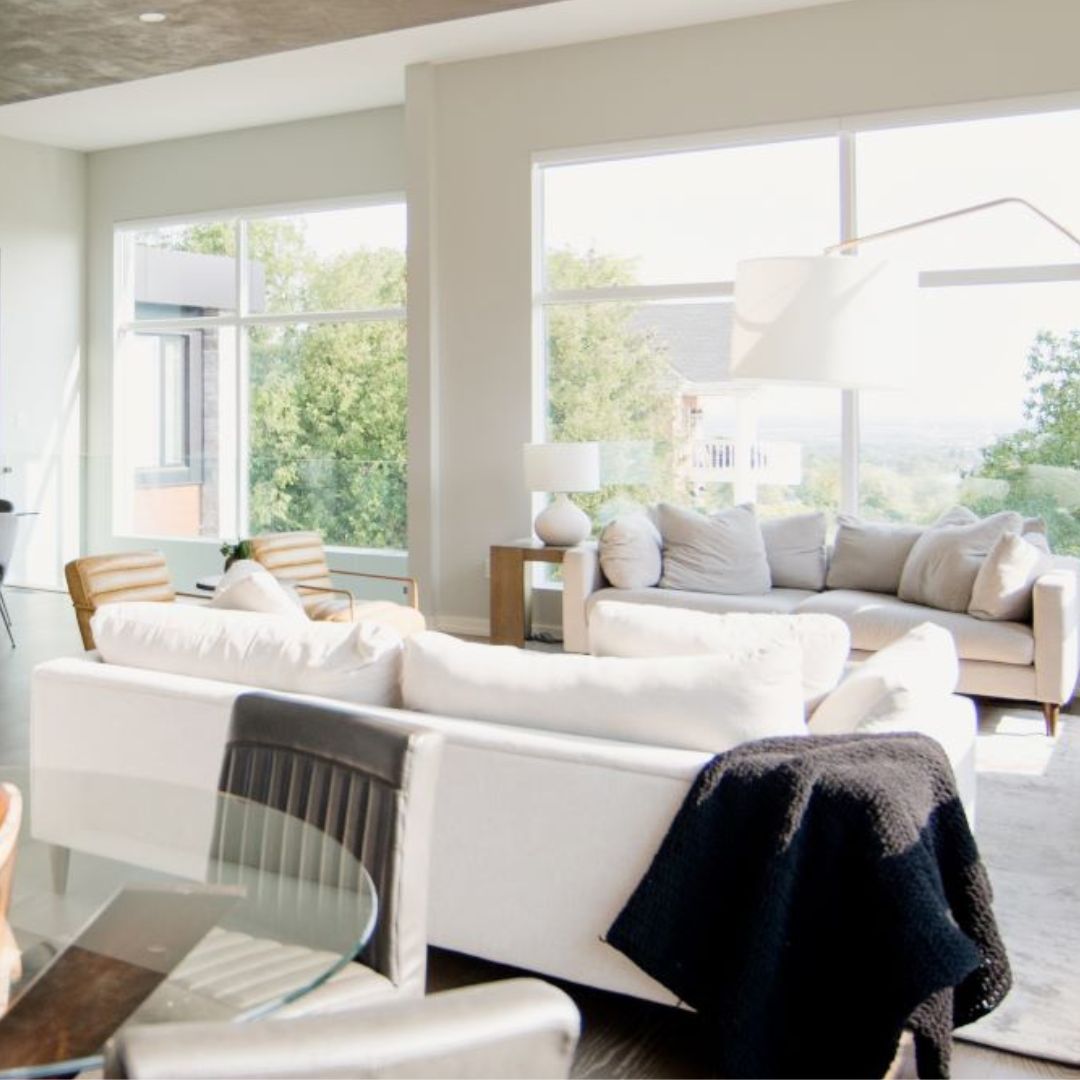 Sunny living room with lots of white couches and big windows. You can see trees and other houses on a hill outside. The room has wooden chairs too, and it looks really open and bright. It's a perfect place to relax and enjoy the view.