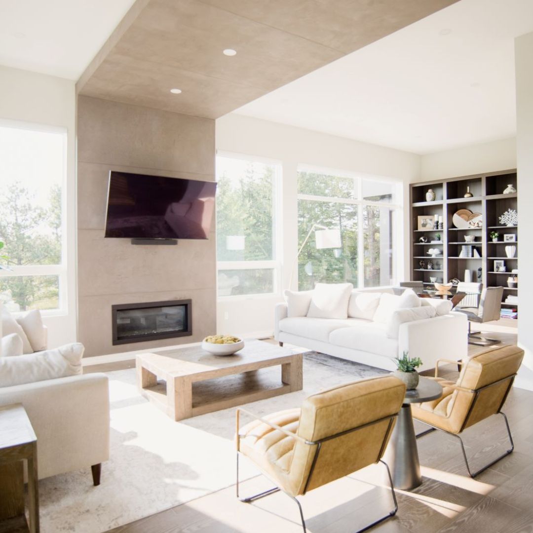 Luxurious modern living room design: bright open-concept space with floor-to-ceiling windows, showing nature views. Features sleek white sofa, stylish tan leather accent chairs, and minimalist wooden coffee table. Wall-mounted TV above linear fireplace creates entertainment focal point. Custom built-in bookshelves offer chic storage. Recessed lighting and natural sunlight blend for perfect ambiance. Ideal inspiration for contemporary home interiors, seamless indoor-outdoor living, and high end residentia