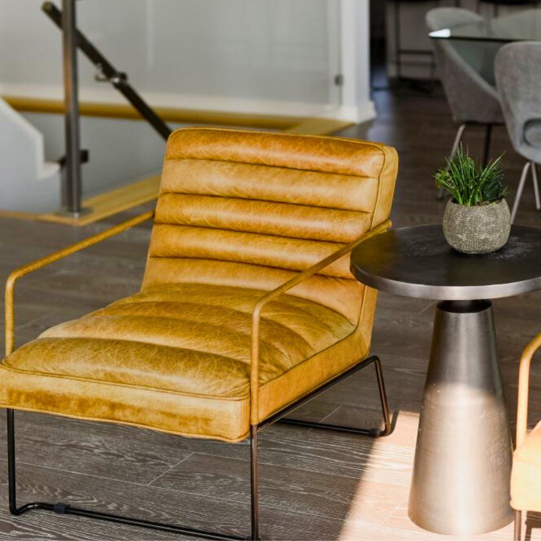 Trendy mustard yellow leather lounge chair with ribbed cushions and sleek metal frame, placed next to a modern side table in a contemporary living space. The chair's warm color pops against dark wood flooring, creating an inviting seating area.