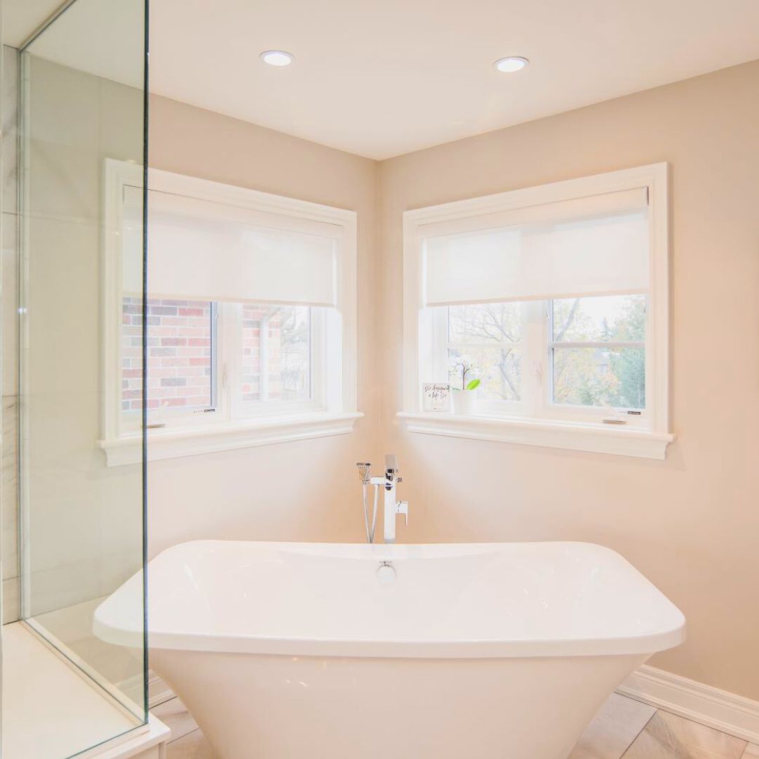 Bright, modern bathroom featuring a freestanding white tub, glass shower enclosure, and large windows. Beige walls and recessed lighting create a spa-like atmosphere.