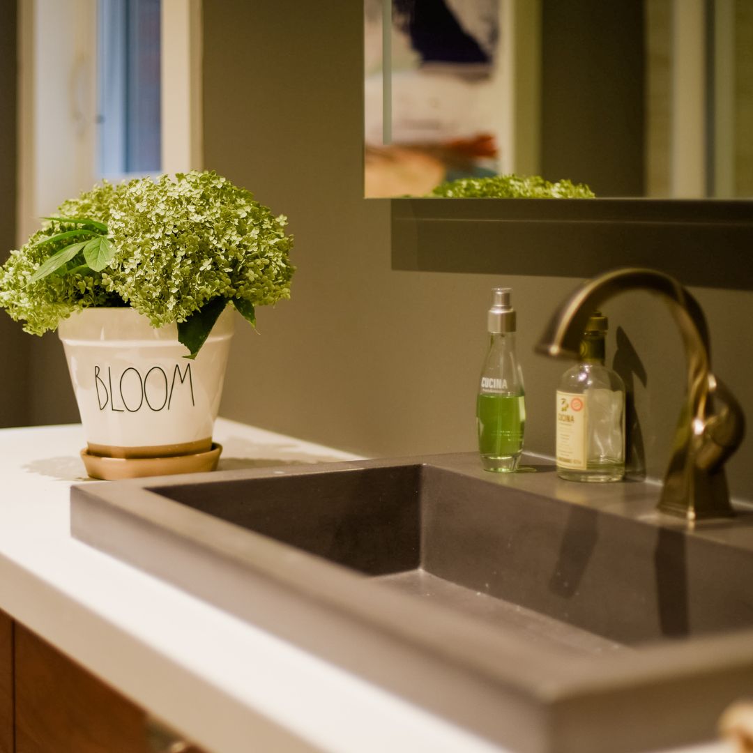 Modern bathroom sink with a matte black basin, brass fixtures, and contemporary decor, including a 'Bloom' plant arrangement. 