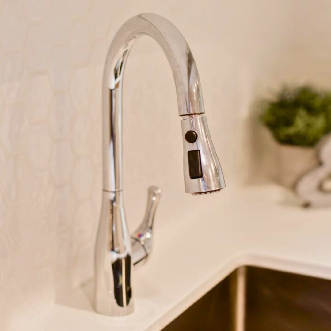 Stylish chrome kitchen faucet with pull-down sprayer. White quartz countertop and beige backsplash in background. Small potted plant adds a touch of green
