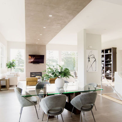 Contemporary dining area featuring a glass table with gray chairs, set against a backdrop of white walls and large windows. A striking concrete ceiling detail runs down the center, adding visual interest. 