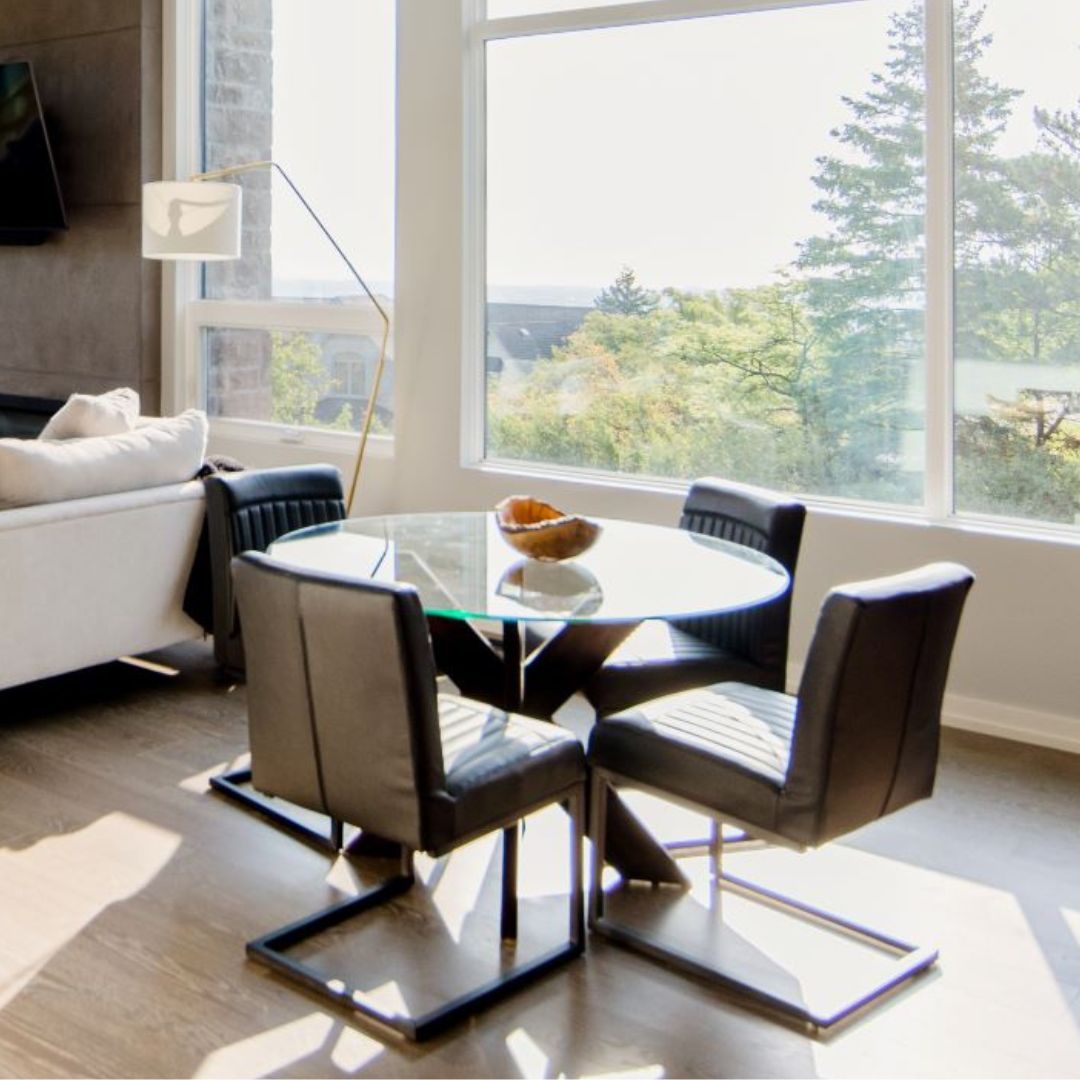 Stylish dining area with round glass table and black chairs. Large windows offer stunning tree views. White sofa and floor lamp visible, creating an airy, contemporary living space.