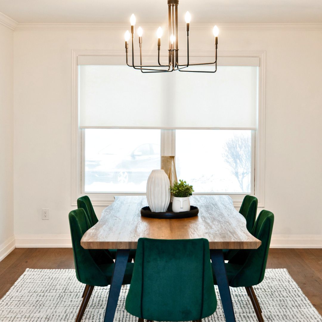 Modern dining room design featuring a rustic wooden dining table paired with elegant green upholstered chairs. Includes a stylish chandelier, neutral decor accents, and large windows