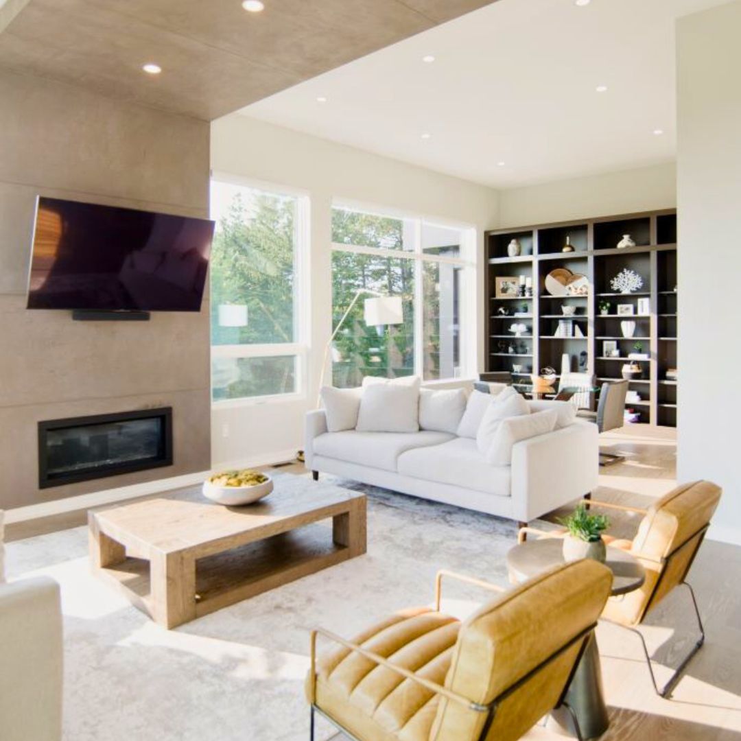 Stunning modern living room design with floor-to-ceiling windows, showcasing natural light and lush outdoor views. Features sleek white sofa, contemporary tan leather chairs, and minimalist wooden coffee table. Focal point includes wall-mounted TV above linear fireplace, perfect for entertainment. Built-in bookshelves add storage and style