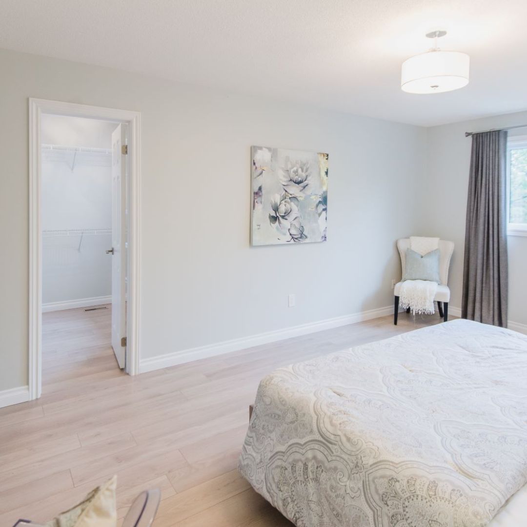 Spacious master bedroom with walk-in closet, light hardwood floors, drum pendant light, floral wall art, and accent chair, displaying modern and airy design