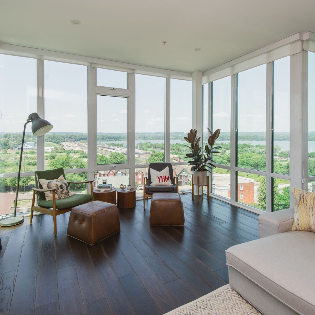 Modern penthouse living room in Hamilton, Ontario featuring floor-to-ceiling windows with panoramic views, a mix of mid-century furniture, and dark hardwood flooring. The design includes green and leather accent chairs, a reading nook, and a neutral color palette.