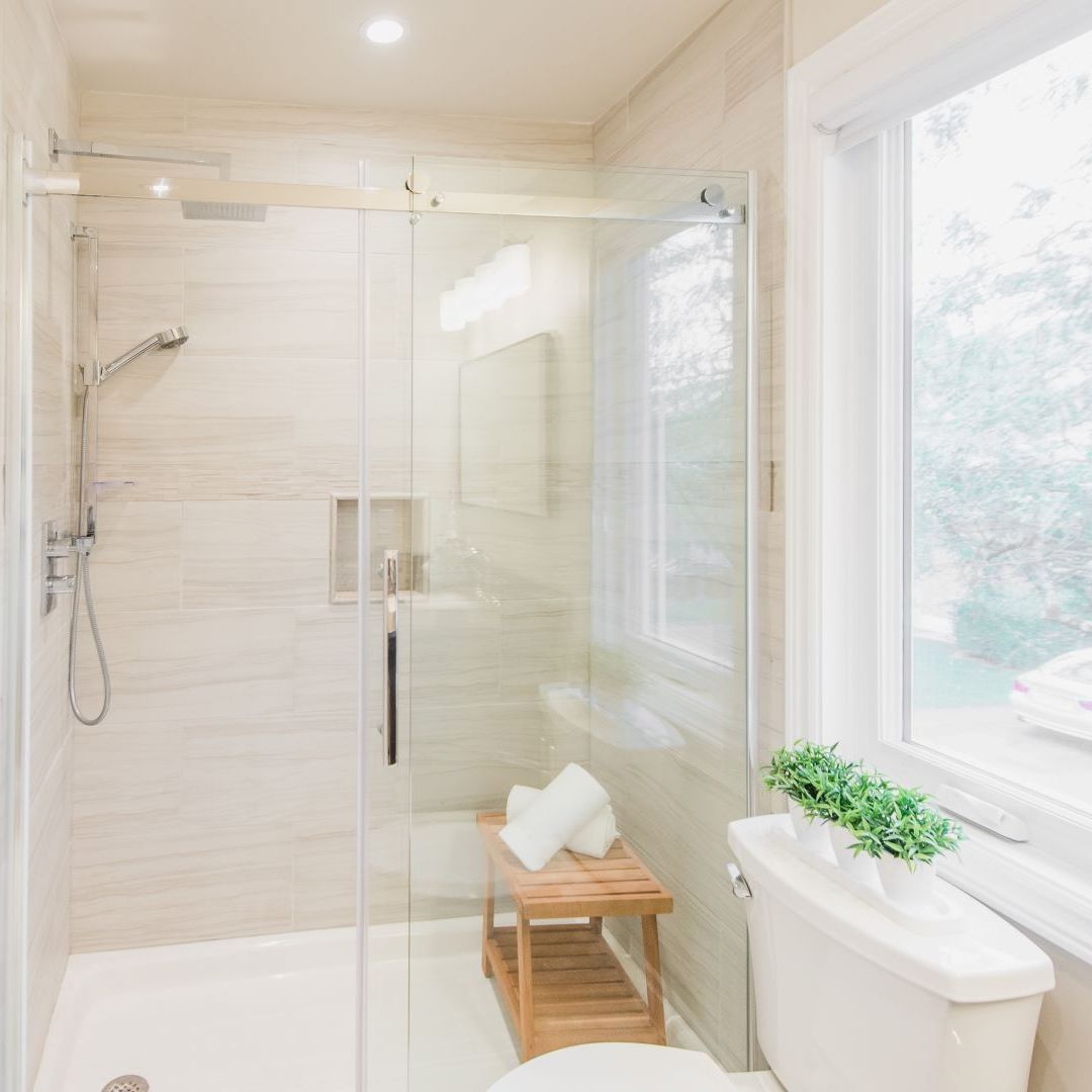 Bright, spacious shower with glass sliding doors, light wood bench, and rainfall showerhead. Large window lets in natural light.