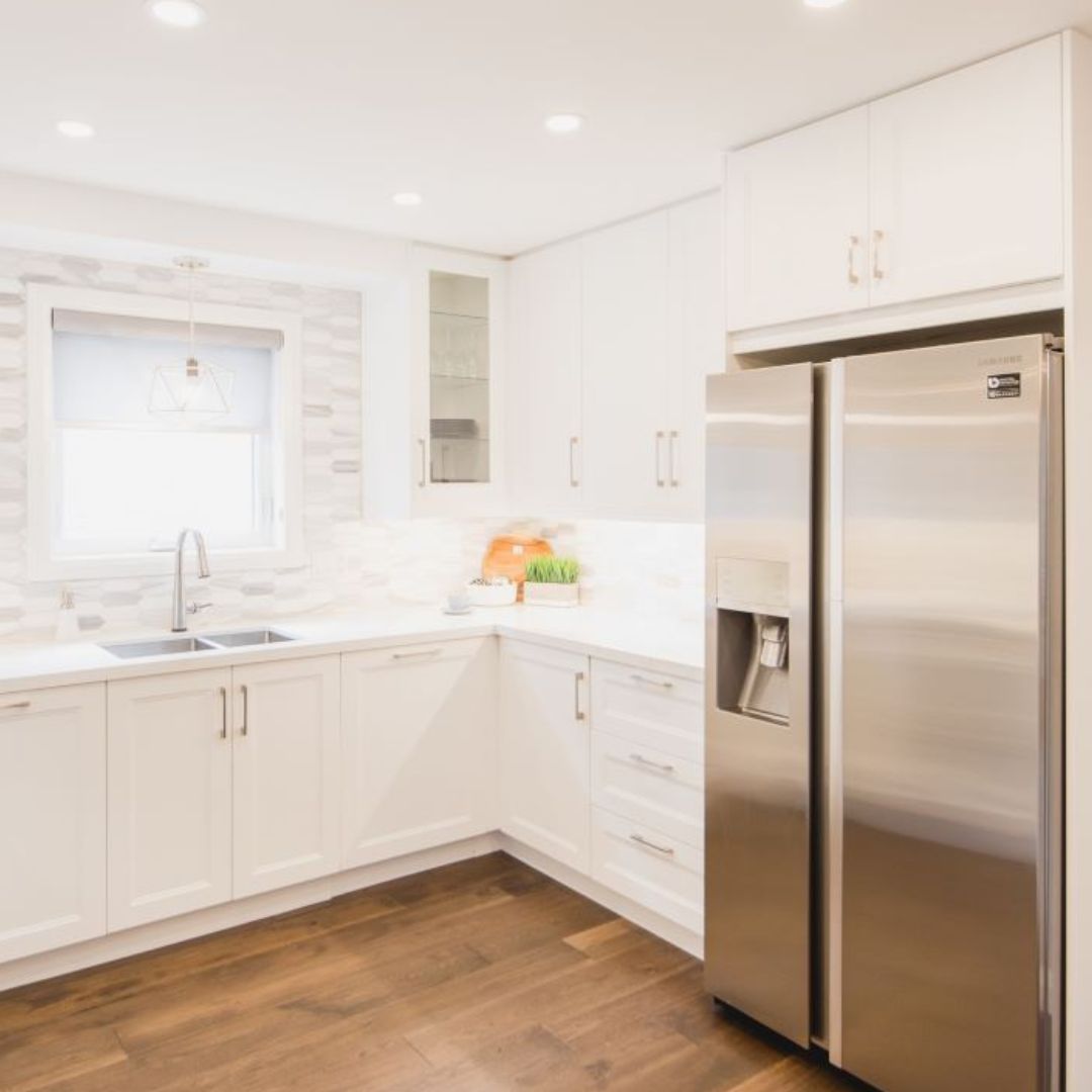 Modern white kitchen with L-shaped layout, shaker cabinets, stainless steel appliances, and warm wood flooring. 