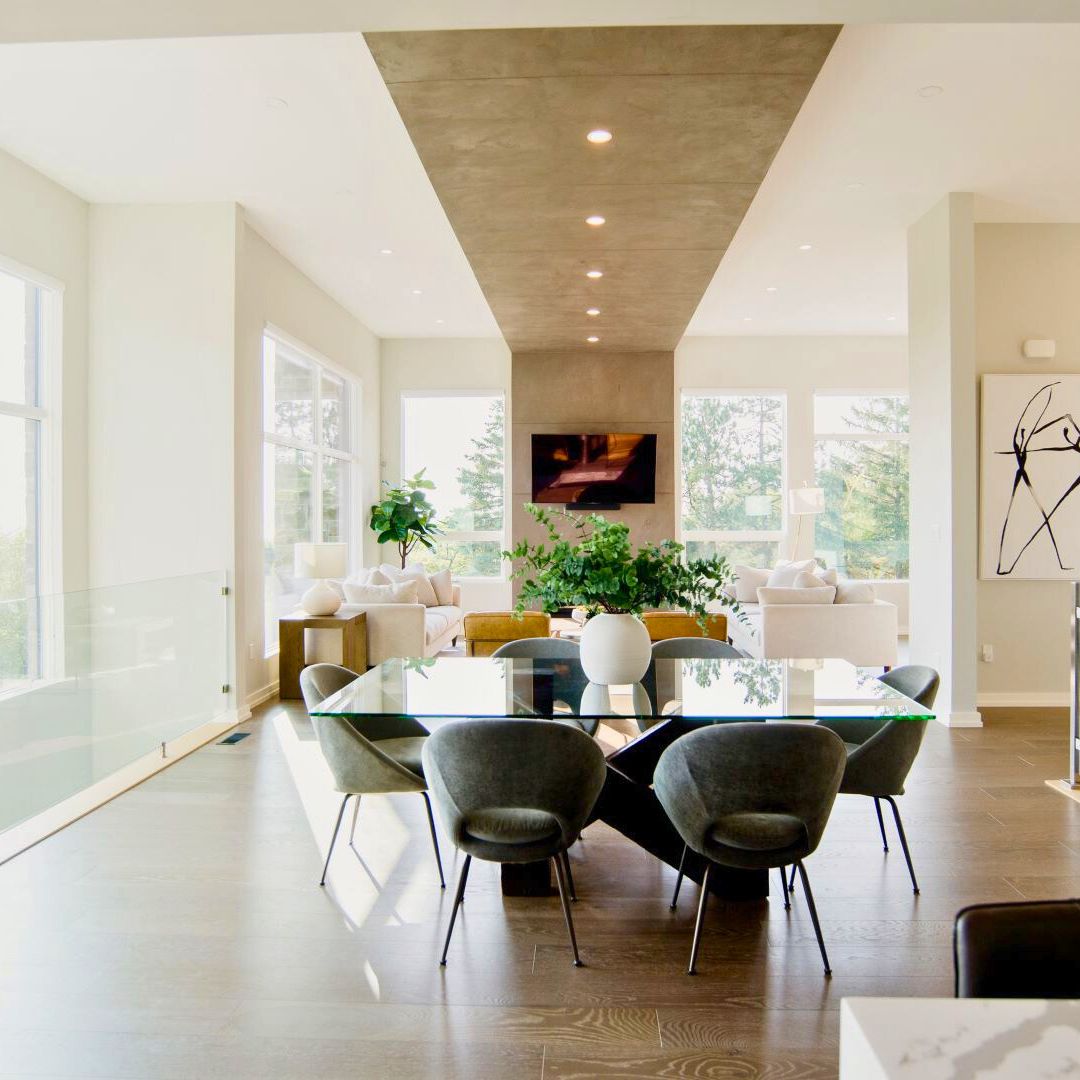 Bright open-concept living and dining area with high ceilings and large windows. A glass dining table with gray chairs sits in the foreground, while a cozy living room with fireplace and TV is visible in the background. Natural light floods the space, highlighting the modern, airy design.