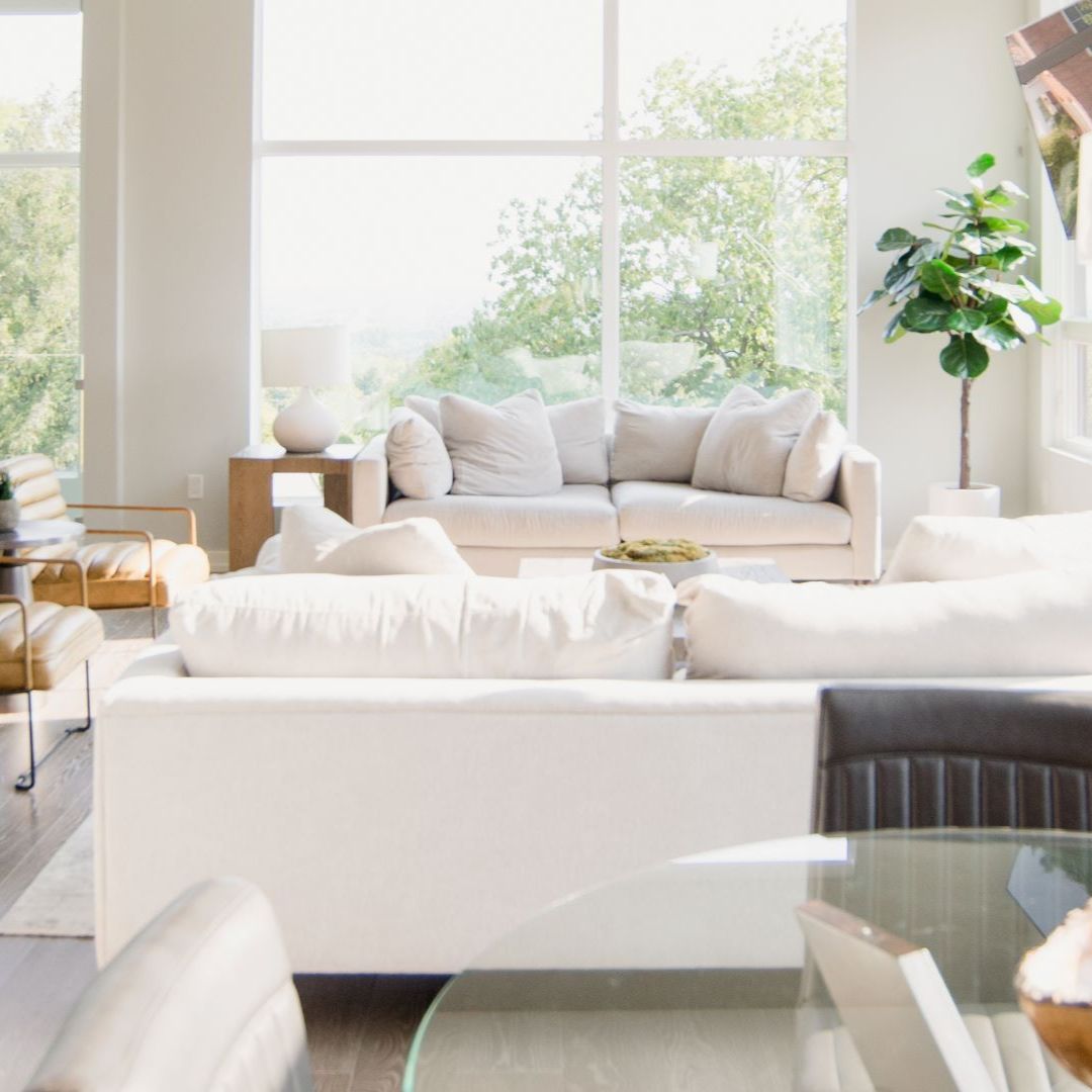 A peaceful coastal living room with comfy white sofas and large windows that show beautiful green scenery. Natural light fills the modern space, which has a fiddle-leaf fig plant and a glass coffee table. It's the perfect mix of indoor and outdoor living, great for luxury home design ideas.