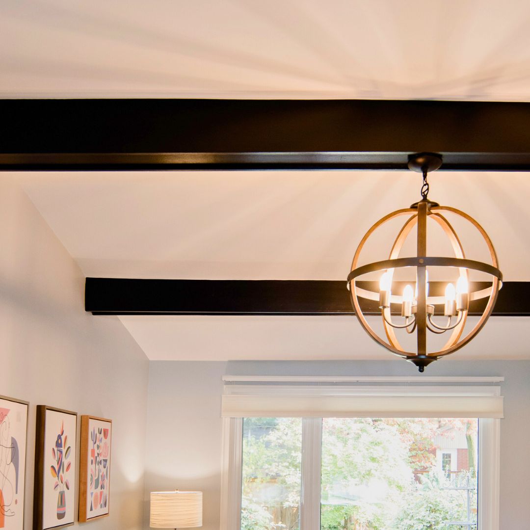 home interior with vaulted white ceiling, dark exposed wood beams, brass spherical pendant light, and large windows offering lush outdoor views