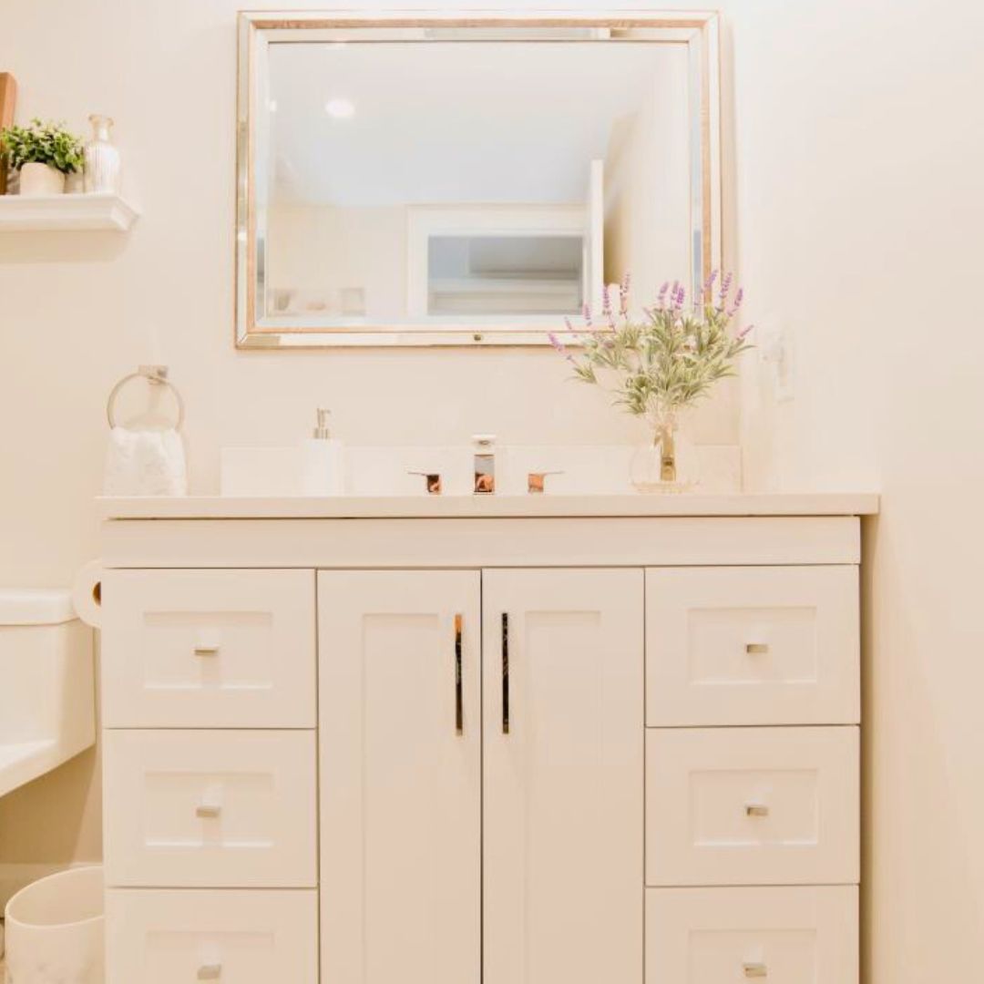 Bathroom vanity with white cabinets, quartz countertop, and rectangular sink. Gold-framed mirror and hardware add elegance. Lavender flowers in vase for a pop of color